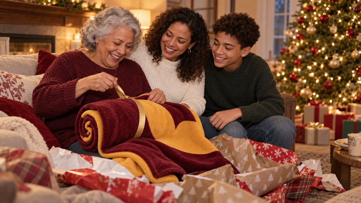 A grandmother unwraps a rolled maroon and gold fleece blanket tied with ribbon beside a decorated holiday tree while her family watches.